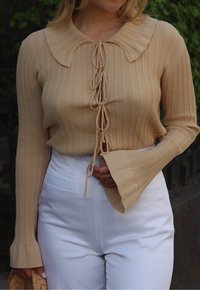 Beige ribbeltruitje met een grote kraag, voorzien van een strikdetail aan de voorkant en uitlopende mouwen, gecombineerd met witte high-waisted broek.