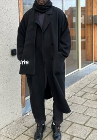 Casaco de lã oversized preto com colarinho de xale, com botões duplos, combinado com um cachecol preto texturizado e calças pretas. Sapatos pretos completam o visual.