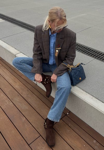 Mujer rubia con gafas, blazer a cuadros, camisa azul, vaqueros y botas marrones sentada en un borde de hormigón, ajustándose la bota, con un bolso acolchado azul cerca.