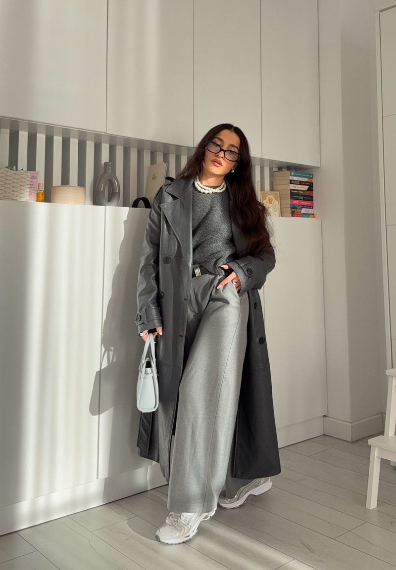 Trench-coat gris, pull gris, pantalon large argenté, baskets blanches et petit sac à main bleu clair. Cheveux longs, lunettes et collier de perles.