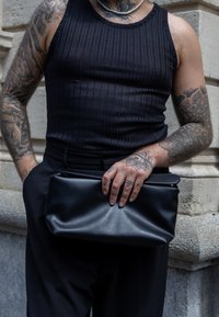 Black leather clutch bag held in hand, featuring a smooth texture and minimalist design, teamed with a ribbed black tank top and tailored trousers.