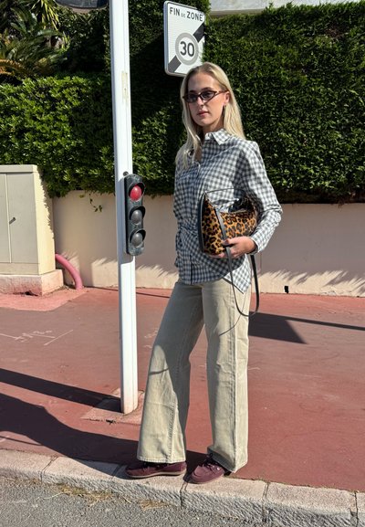 Mujer con gafas de sol, camisa gris de cuadros, pantalones beige, sosteniendo un bolso con estampado de leopardo, parada en la acera junto a un semáforo y una señal de límite de velocidad.