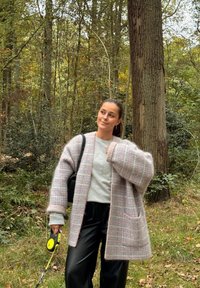 Gros cardigan en plaid rose et gris, duveteux et oversized, avec de grandes poches, associé à un haut gris clair et un pantalon noir brillant, tenu à côté d'une laisse.