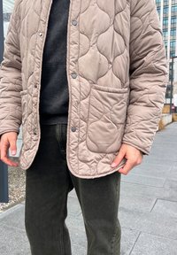 Κοντό μπεζ μπουφάν με κορδέλες και μια θήκη, σε συνδυασμό με σκούρα πράσινα παντελόνια. Το ύφασμα έχει υφή σχεδίου.