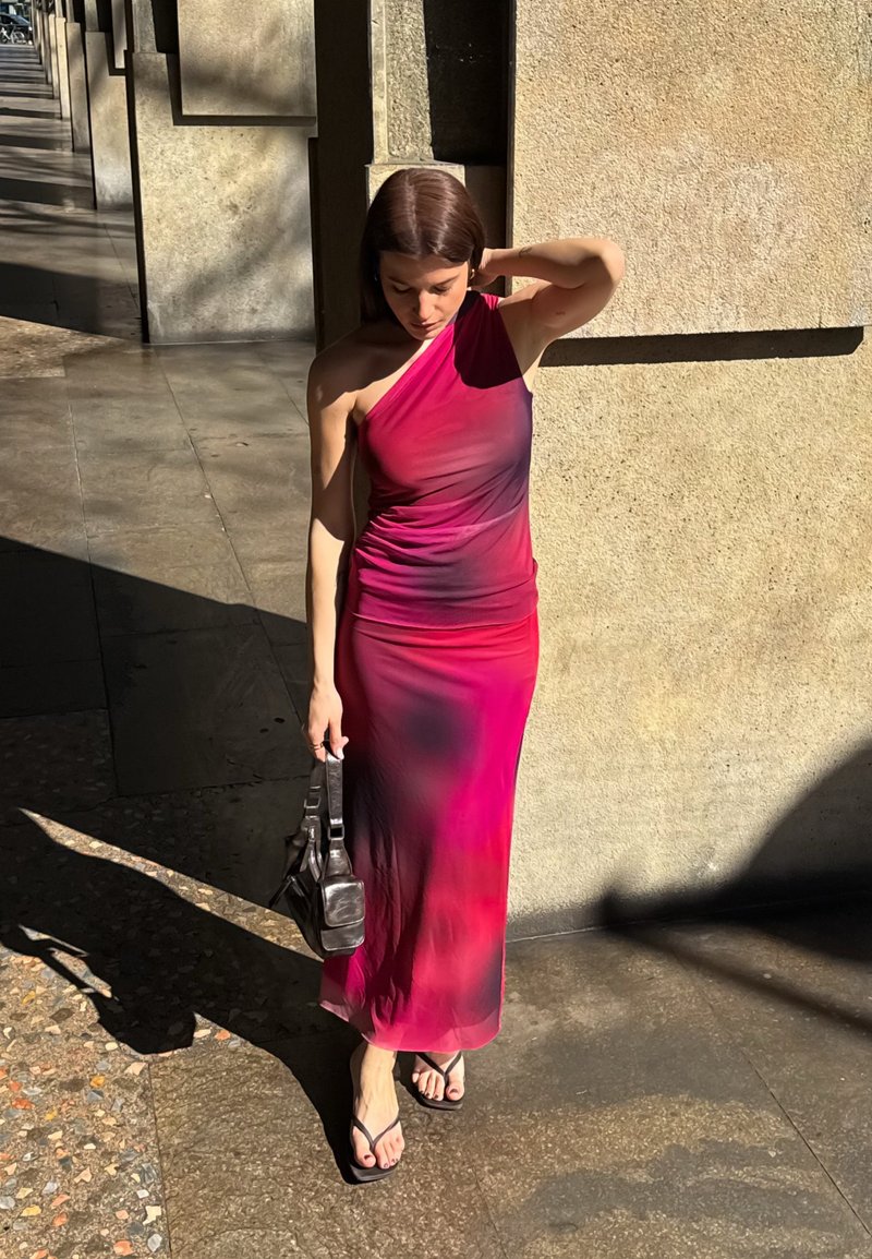 Femme dans une robe rose et violette à une épaule, tenant un sac noir, debout au soleil à côté d'un mur en pierre texturé.