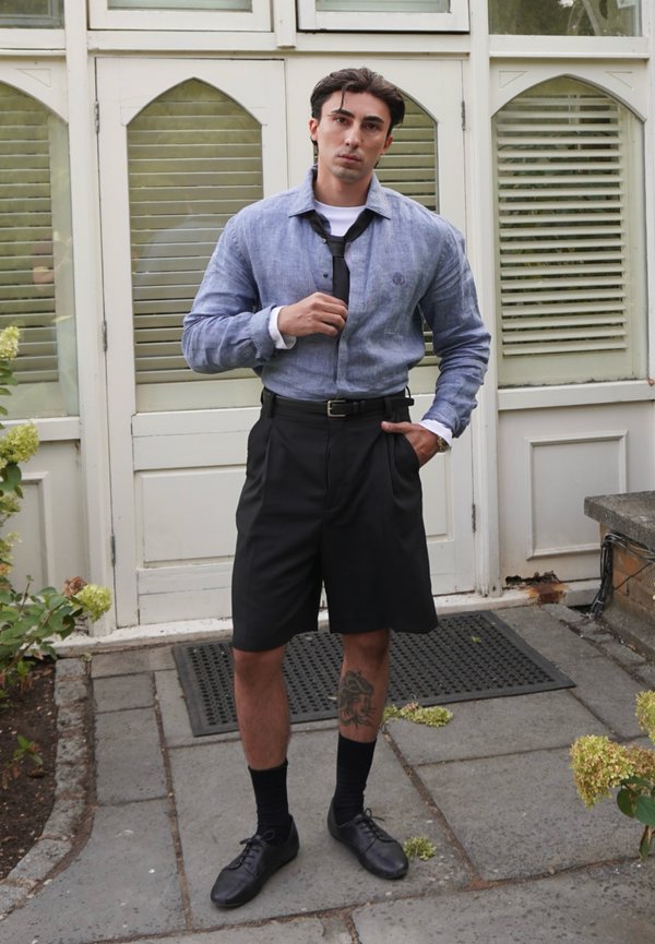 Homme debout dehors devant des portes blanches, portant une chemise bleue, une cravate noire, un short noir, des chaussures habillées et un tatouage sur son mollet gauche.