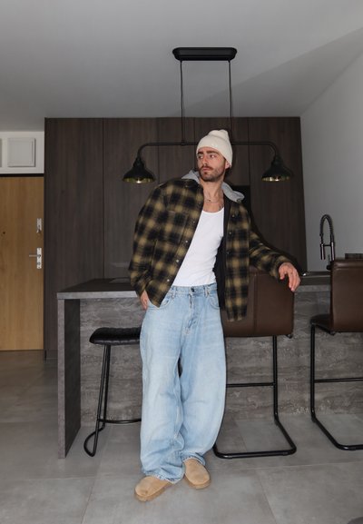 Hombre con gorro blanco, camisa de cuadros, camiseta blanca sin mangas y jeans holgados, apoyado en la encimera de la cocina con taburetes de bar y luces colgantes detrás de él.
