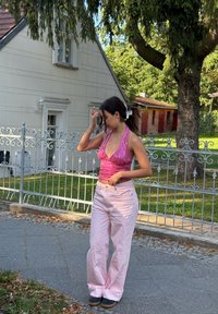Top halter rosa brilhante com lantejoulas, combinado com calças largas rosa claro e ténis escuros, diante de um jardim verde e uma cerca cinza.