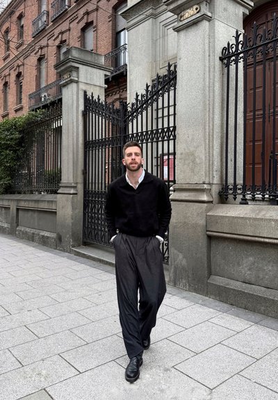 Un suéter negro sobre una camisa blanca con cuello, acompañado de pantalones negros a medida. El fondo presenta un edificio de piedra y puertas de hierro.