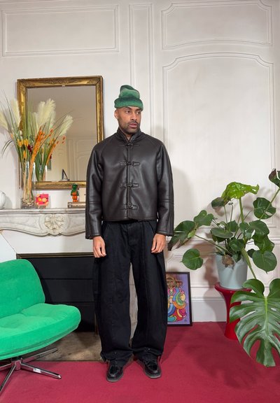 Chaqueta corta de cuero marrón con cierres de pasador, combinada con pantalones negros holgados. Un gorro de punto verde completa el conjunto. Ambiente interior.