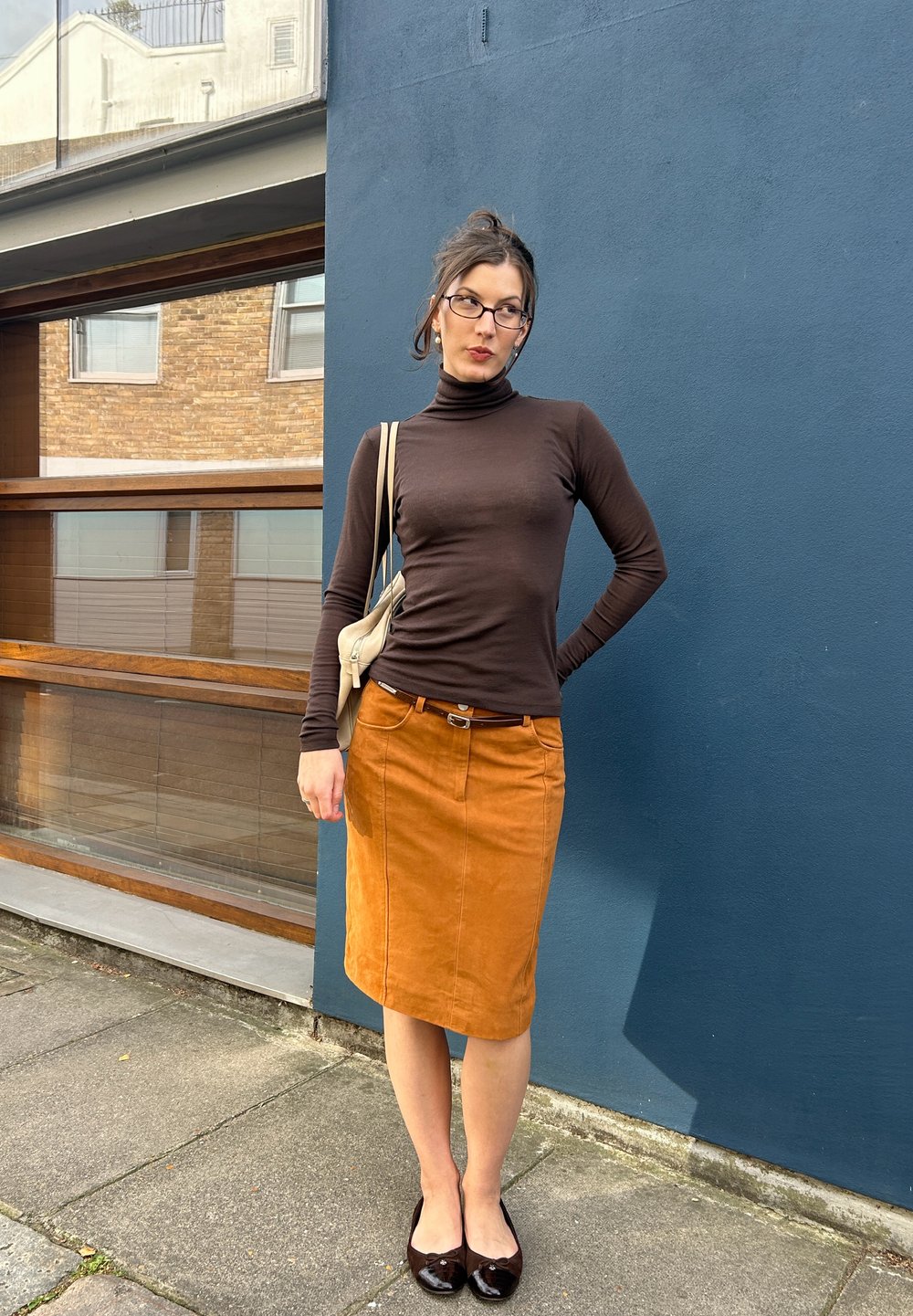 Brun tilpasset turtleneck, oransje suedepensko, svarte lakksko og en beige veske. Stående mot en blå vegg med treaksenter.