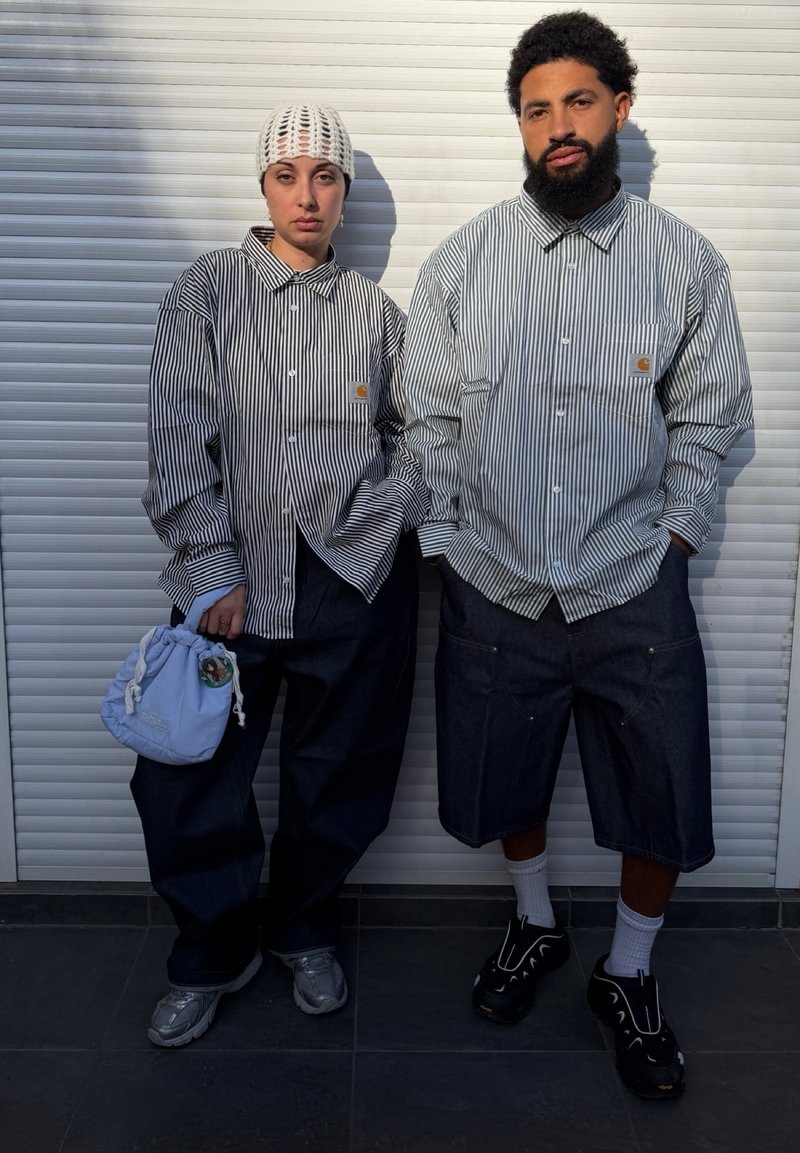 Twee personen dragen zwart-wit gestreepte overhemden met knopen en donkere broeken, staand tegen een witte luikenachtergrond in zacht zonlicht.