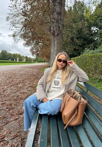 Henkilö istuu vihreällä penkillä puistossa, pukeutuneena ruudulliseen bleiseriin ja vaaleaan neuleeseen, ja heidän vieressään on vaaleanruskea käsilaukku.