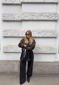 Γυναίκα με μακριά ξανθά μαλλιά που φοράει σκούτερ σε σκούρο χρώμα, φαρδιές μαύρες παντελόνες και κρατά μια μεγάλη μαύρη τσάντα, στέκεται μπροστά σε έναν τοίχο από πέτρα με υφή.