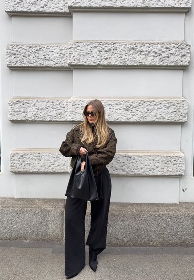 Mujer con cabello rubio largo, vestida con un suéter oscuro de gran tamaño y pantalones anchos negros, sosteniendo un bolso grande negro, de pie frente a una pared de piedra texturizada.
