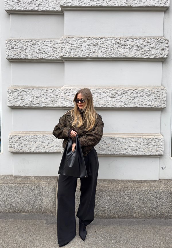 Frau mit langen blonden Haaren, die einen dunklen, übergroßen Pullover und weite schwarze Hosen trägt, hält eine große schwarze Handtasche und steht vor einer strukturierten Steinwand.