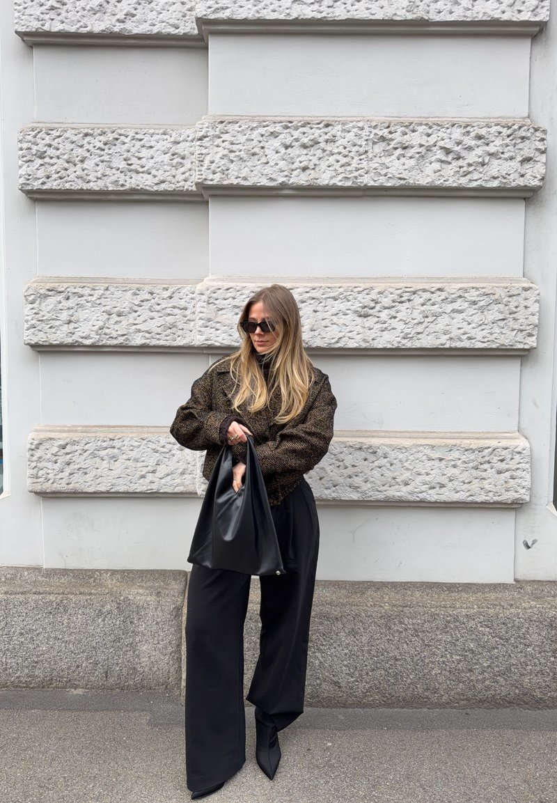Femme aux longs cheveux blonds vêtue d'un pull ample sombre et d'un large pantalon noir, tenant un grand sac à main noir, debout devant un mur de pierre texturé.