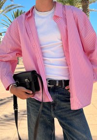 Lichtroze gestreepte button-up shirt gedragen over een witte tanktop. Zwarte tas met zilveren accenten, donkere jeans en een zilveren gesp riem.