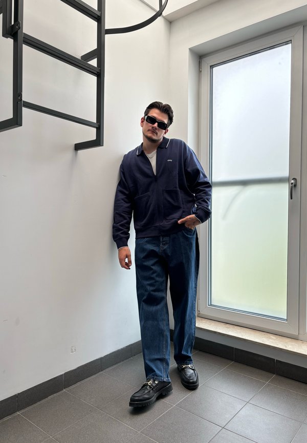 Marinblå zip-up jacka med vit krage, denim jeans och svarta skor med metallaccent. Står mot en vägg nära ett fönster.