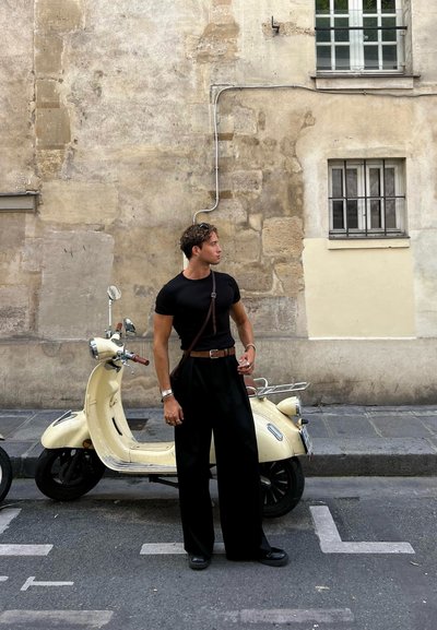 Camiseta negra ajustada, pantalones negros holgados y un bolso bandolera marrón. Scooter vintage amarilla junto a una pared de piedra texturizada.