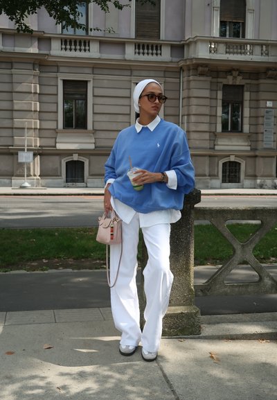 Sudadera de gran tamaño en azul claro con cuello blanco, pantalones de pierna ancha blancos y un pequeño bolso rosa con una correa colorida. Sosteniendo una bebida.