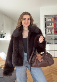Femme portant une veste en fourrure brun foncé tenant un sac à main en cuir marron avec un porte-clés petit ours en peluche, debout dans un salon moderne.