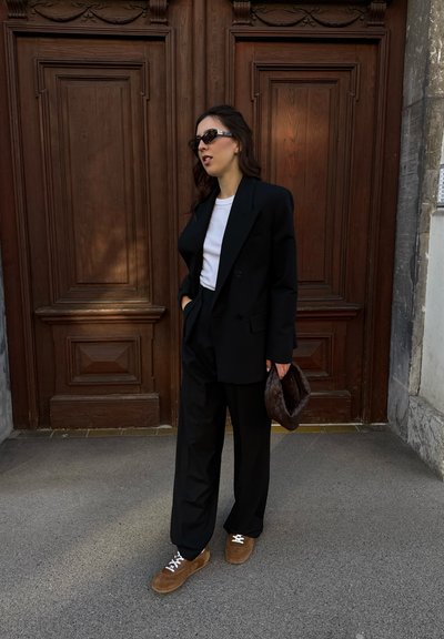 Blazer y pantalones negros a medida sobre una camisa blanca, combinados con zapatillas deportivas marrones y un bolso marrón texturizado. Las gafas de sol completan el look.
