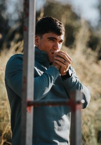 Veste polaire bleue à fermeture éclair, coupe ajustée, manches longues. Mains jointes pour se réchauffer, tatouage visible au poignet, arrière-plan naturel flou.