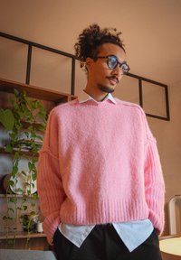 Homme aux cheveux bouclés attachés, portant des lunettes et un pull rose par-dessus une chemise blanche, debout à l'intérieur près d'une plante et d'une étagère en bois.