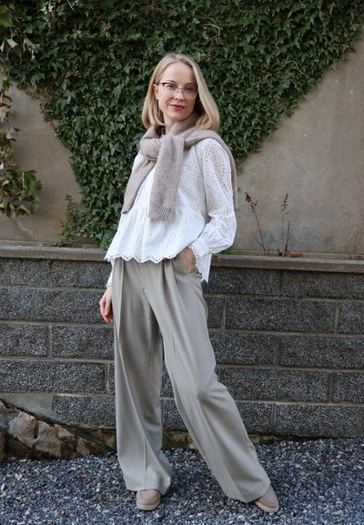 Mujer con gafas que lleva una blusa blanca con ojales, pantalones anchos beige y una bufanda topo, de pie al aire libre frente a una pared cubierta de hiedra.