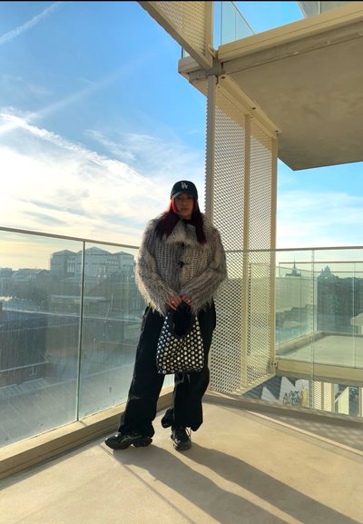 Suéter gris y difuso con flecos, pantalones cargo negros y un bolso negro con tachuelas. Combinado con una gorra negra y zapatillas oscuras.