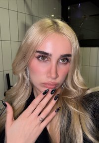 Jeune femme aux longs cheveux blonds montrant des ongles noirs en forme d'amande avec des points blancs et un petit tatouage en forme de cœur sur le doigt, portant des boucles d'oreilles pendantes.