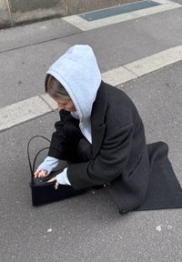 Giovane in un cappotto nero e felpa grigia accovacciato sul marciapiede, che apre o chiude una borsa nera.