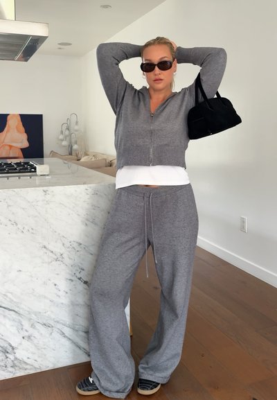 Mujer con conjunto gris de descanso y gafas de sol sostiene un bolso negro sobre el hombro, de pie en una cocina moderna con isla de mármol y suelo de madera.