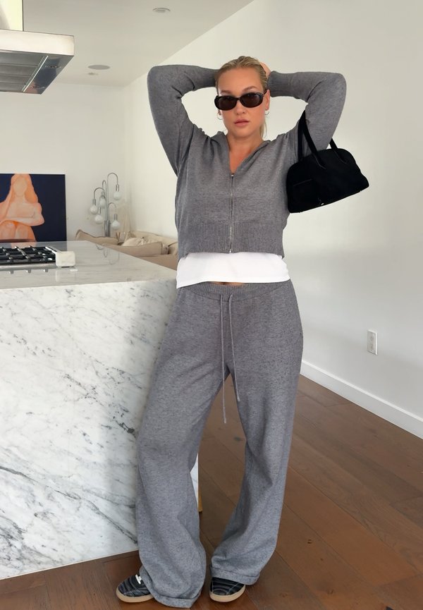 Femme vêtue d'une tenue grise de détente et portant des lunettes de soleil, tenant un sac à main noir sur l'épaule, debout dans une cuisine moderne avec un îlot en marbre et un sol en bois.