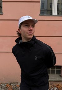 Zwarte lichte jas met hoge kraag, met grijze accenten en logo. Witte pet met logo. Achtergrond: roze muur.