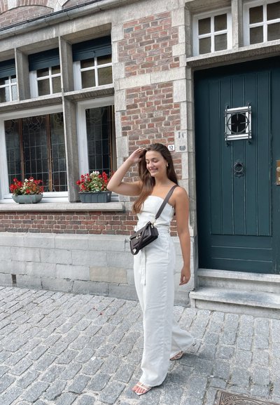 Mono blanco con un corpiño ajustado y pantalones anchos, combinado con un pequeño bolso cruzado negro. Calle de adoquines y pared de ladrillo con macetas de flores.