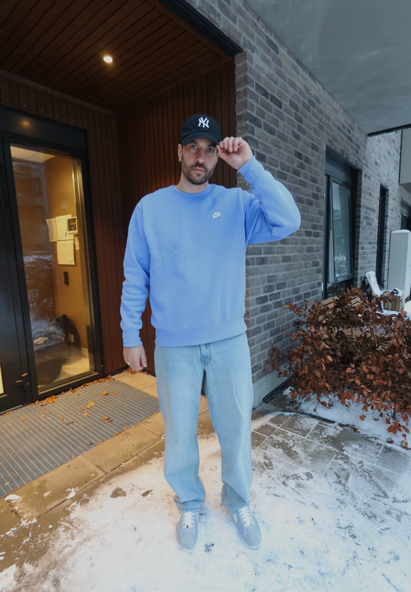 Homme en sweat-shirt Nike bleu clair, jean et baskets grises ajustant une casquette noire, debout sur un sol enneigé près de l'entrée d'un bâtiment en briques.