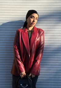 Blazer in pelle rossa con spalle strutturate, maniche lunghe e dettagli con bottoni, indossato sopra una maglia nera strutturata, accessoriato con una piccola borsa nera.