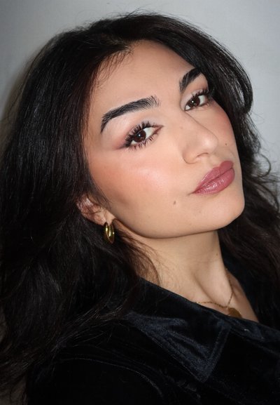 Persona con cabello largo y oscuro, lleva pendientes de aro dorados y una blusa de terciopelo negro, con maquillaje suave y un color de labios neutro.