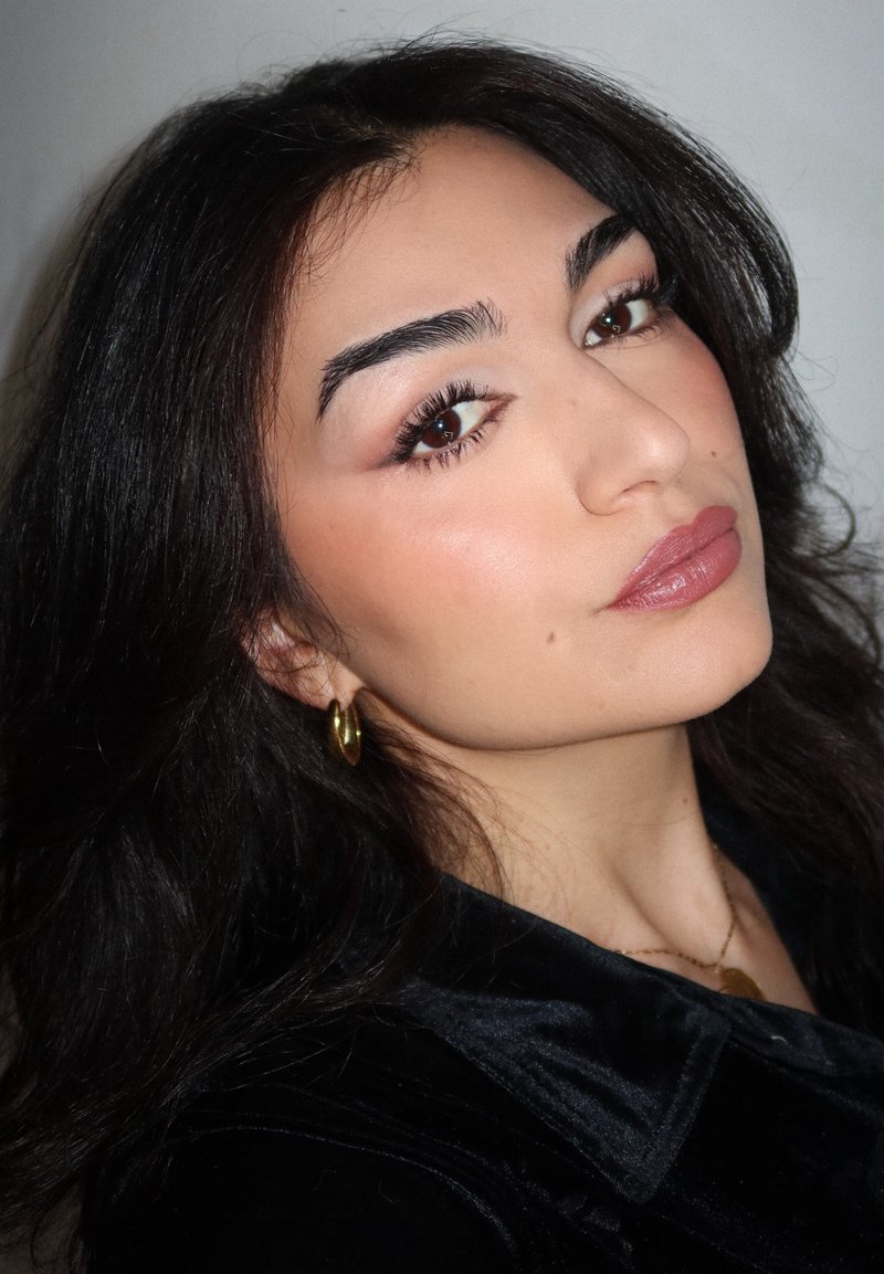 Personne aux cheveux longs et foncés, portant des boucles d'oreilles en or en forme de créoles et un haut en velours noir, avec un maquillage doux et une couleur de lèvres neutre.