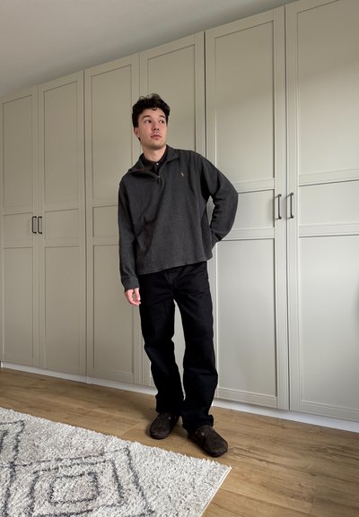 Pulóver de forro polar de carbón con cuello y cremallera de un cuarto, combinado con pantalones negros y zapatos marrón oscuro, con un fondo de gabinete claro.