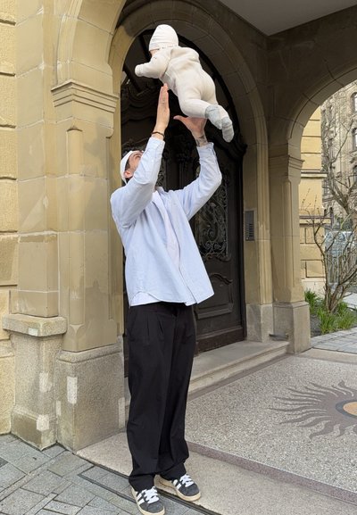 Adulto con camisa azul claro y pantalones negros levantando a un bebé vestido de blanco cerca de la entrada de un edificio con arcos de piedra.