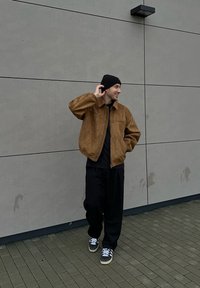 Brown suede bomber jacket, black t-shirt, loose black trousers, black beanie, and grey trainers with white laces. Standing against a grey wall.