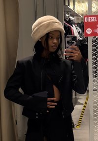 Μαύρο κοντό μπουφάν με ψηλό γιακά και έντονα μανίκια, συνδυασμένο με ένα κρεμ γούνινο καπέλο. Η αντανάκλαση στον καθρέφτη αποτυπώνει το πάνω μέρος του σώματος.