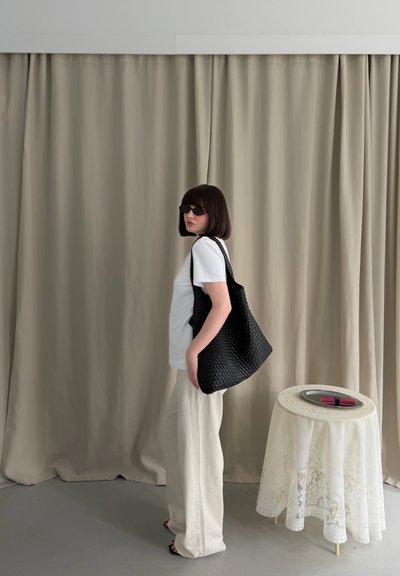 Mujer con el cabello corto y oscuro y gafas de sol, de pie sosteniendo una gran bolsa negra tejida, cerca de una mesa pequeña con mantel de encaje y un plato con objetos.