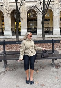 Beige trenssitakki, jossa korkea kaulus, mustat lyhyet housut ja pyöreäkärkiset mustat kengät. Istuu puiston penkillä, ympärillä pudonneita lehtiä.