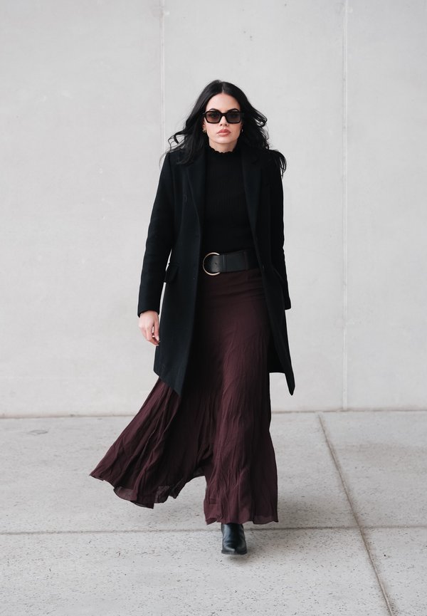 Long burgundy skirt with a flowing texture, black fitted top, black coat, and ankle boots. Accessories include round sunglasses and a belt.