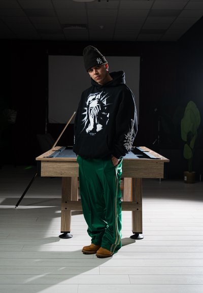 Joven con sudadera negra con capucha y gorro, pantalones deportivos verdes y botas marrones, de pie frente a una mesa de billar en una habitación con poca luz.