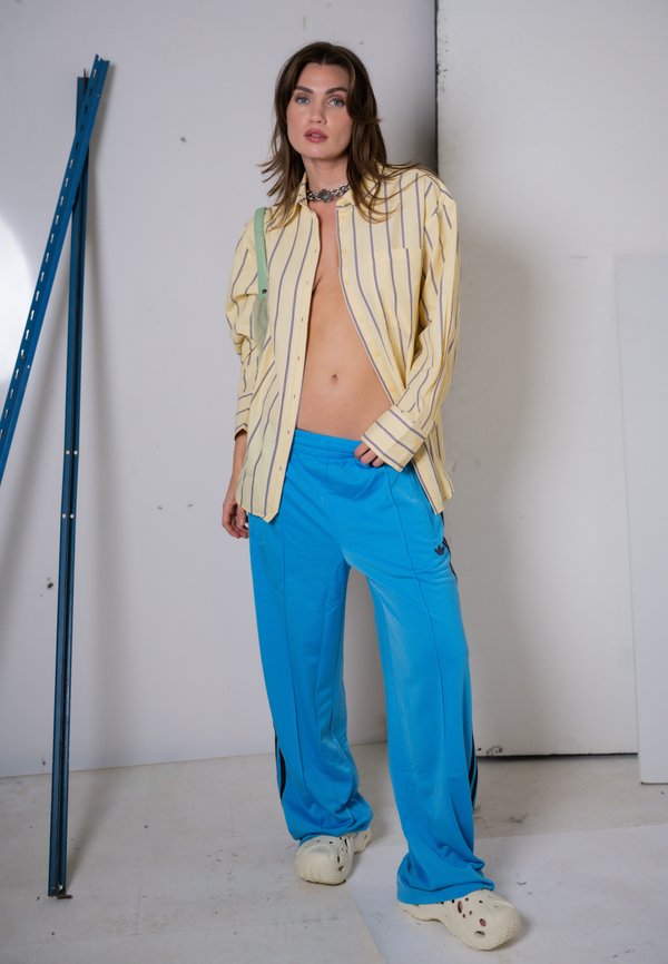 Femme portant une chemise rayée jaune ouverte, un pantalon bleu vif, des chaussures blanches sans lacets et un collier en chaîne, debout devant un fond blanc uni.